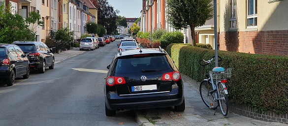 Eine Straße auf der links und recht aufgesetzt geparkte Autos stehen. Sehr enge bis nicht vorhandene Gehwegbreiten. 